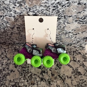 May include: A pair of novelty earrings featuring miniature monster trucks. The trucks have black and purple bodies with green wheels. The text "Grave Digger" is visible on the top of the trucks. The earrings hang from silver-coloured hooks.