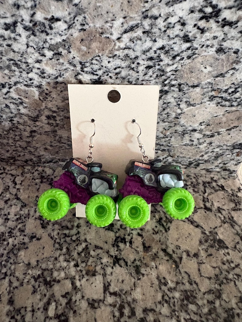 May include: A pair of novelty earrings featuring miniature monster trucks. The trucks have black and purple bodies with green wheels. The text "Grave Digger" is visible on the top of the trucks. The earrings hang from silver-coloured hooks.