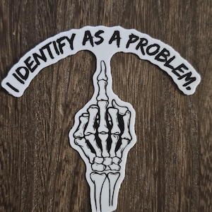 Puede incluir: Una pegatina blanca con una mano de esqueleto negra haciendo el dedo medio. La pegatina tiene el texto "Me identifico como un problema." en un arco negro sobre la mano.