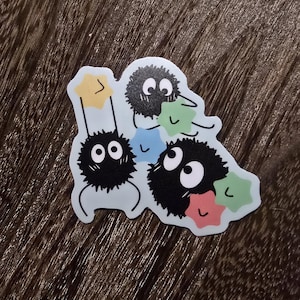 May include: A sticker featuring three black fuzzy creatures with large eyes, each holding a star-shaped object in different colours: blue, green, and pink. The creatures are on a light blue background.