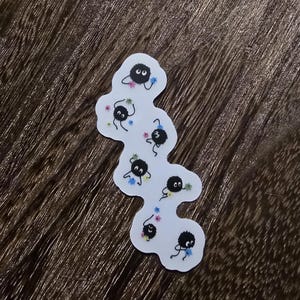 May include: A sticker featuring a group of black, fluffy, cartoon creatures with large eyes. The creatures are surrounded by small, colourful flowers.