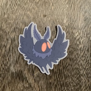 May include: A sticker featuring a stylized moth-like creature with a dark blue body, large wings, and orange eyes. The wings have a feathery texture, and the creature has a round head. The sticker is on a wooden surface.