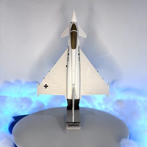 May include: A white Eurofighter Typhoon model airplane with black accents and a black cross symbol on the wing. The model is mounted on a gray stand with the text "Eurofighter Typhoon".