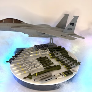 May include: A gray scale model of an F-15 fighter jet with the tail markings PB 1994, mounted on a stand. Below the aircraft, a circular display holds an assortment of miniature gray missiles and olive green bombs arranged in rows on a white surface.