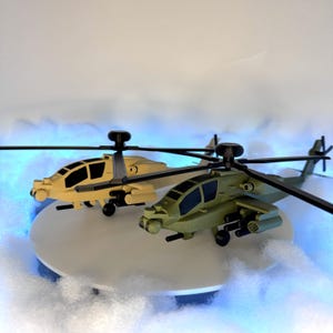 May include: Two toy helicopters, one tan and one olive green, are displayed on a white surface. The helicopters have black rotors and windows. The background features a blue glow and white fluffy material, creating a playful scene.