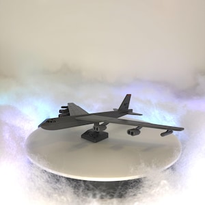May include: A gray B-52 bomber model with detailed wings, engines, and tail. The model sits on a white platform, with the text "B-52" visible. The background features a misty, atmospheric effect, enhancing the model's presentation.