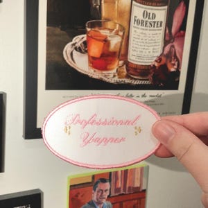 Könnte beinhalten: Ein bestickter, weißer, ovaler Aufnäher mit rosa Einfassung und dem Text "Professional Yapper" in rosa Schrift. Der Aufnäher hat kleine goldene Blumenakzente. Der Hintergrund umfasst gerahmte Kunstwerke und eine Hand, die den Aufnäher hält.