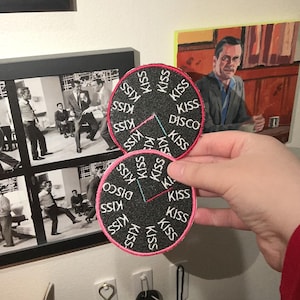 May include: Two circular black patches with the words "KISS DISCO" and "KISS" repeated around the edges. The patches have a pink border and a clock-like design with teal and red hands. The patches are held up in front of a wall with framed pictures.