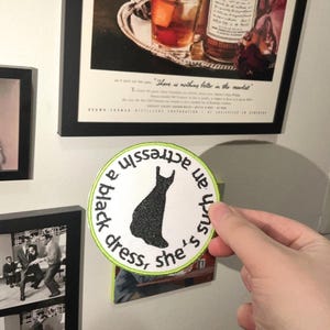 May include: A round, white patch with the text "she's such an actress in a black dress" in a circular pattern. A black dress silhouette is in the center, outlined with a green border. The patch is held up by a hand.