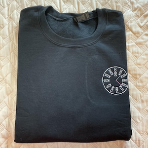 May include: Black crewneck sweatshirt with a circular graphic on the lower right. The graphic features the words "DISCO KISS" and "SSTK KISS" in a circle, with a pink line segment. The sweatshirt is folded.