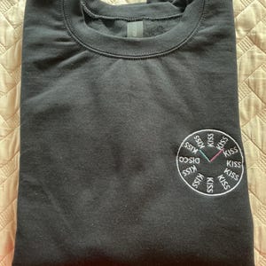 May include: A black crew neck sweatshirt with a circular embroidered design on the left chest. The design features the word "DISCO" and the word "KISS" repeated around a clock face with pink, green, and black hands.