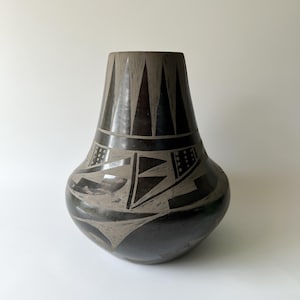 May include: A black and gray ceramic vase with a rounded body and a flared neck. The vase features geometric designs, including triangles and angular shapes, in a contrasting gray color. The vase has a smooth, matte finish.