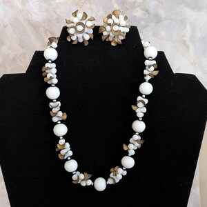 May include: A white and gold necklace and earrings set. The necklace is made of white beads with gold accents. The earrings are small and have a flower shape.