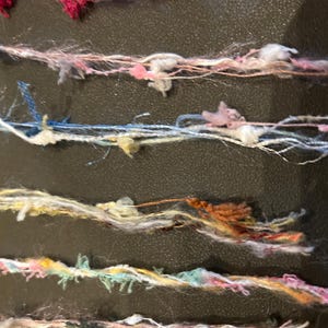 May include: Close-up of several strands of colorful yarn. The yarn strands vary in color, including pink, blue, yellow, and white, with textured, knotted sections. The yarn appears to be a mix of textures and thicknesses, suitable for various craft projects.