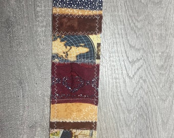 May include: A vertical strip of fabric made from different patterned pieces of fabric. The fabric strip is made up of a white lace, a blue fabric with white dots, a brown fabric with a zig-zag pattern, a brown fabric with a globe pattern, a burgundy fabric with a sparkly pattern, a beige fabric with a floral pattern, a beige fabric with a map pattern, and a burgundy fabric with a zig-zag pattern.