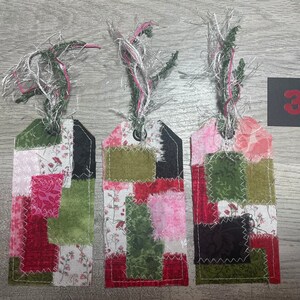 May include: Three fabric gift tags with a patchwork design in shades of pink, red, green, and black. Each tag has a frayed string loop at the top and a hole for a ribbon. The tags are numbered "3" on a black square.