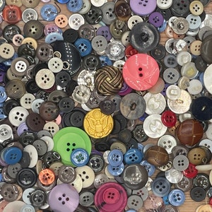 May include: A collection of various buttons in different sizes, colors, and materials. The buttons include shades of black, white, gray, pink, green, and blue. Some buttons have four holes, while others have two. There are also buttons with decorative designs.