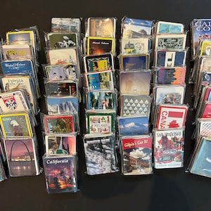 May include: A collection of over 50 colorful souvenir magnets featuring various locations and landmarks. The magnets are arranged in rows and are mostly rectangular in shape. Some of the magnets feature images of cities, states, countries, and landmarks, while others feature text or logos.