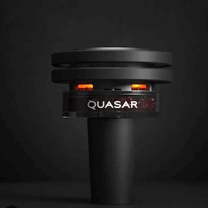 May include: A black device with the word "QUASAR" in white lettering. The device has a cylindrical base and a layered top with a glowing orange interior. The background is a dark, solid color.