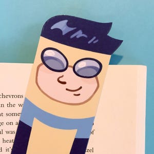 Puede incluir: Un marcapáginas de estilo caricatura con un personaje con gafas, cabello oscuro y piel clara. El marcapáginas es principalmente azul marino, con una cara amarilla clara y una camisa azul. Está colocado en un libro.