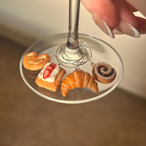 May include: A wine glass with a silver stem, holding a set of miniature food charms. The charms include a pretzel, a pastry with strawberries, a croissant, and a cinnamon roll. The charms are attached to the glass with silver wire.