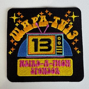 Pode incluir: Um patch quadrado preto com texto e gráficos bordados. O patch apresenta as palavras "TV PARTY" em laranja, uma televisão retrô com o número "13" e o texto "MUM-A-THON SPONSOR" em rosa.