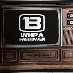 May include: A vintage television set with the WHPA Fairhaven logo displayed on the screen. The image is flanked by color bars and portraits. The television has a wooden cabinet with knobs and a speaker.