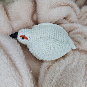 May include: A white crocheted bird with an orange beak and black eyes. The bird is lying on a soft, fluffy, light pink blanket.