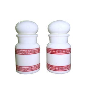 May include: Two white milk glass apothecary jars with rounded lids. Each jar features a cylindrical body decorated with two horizontal bands of intricate red geometric patterns. The jars have a smooth, opaque finish and a classic pharmacy design.