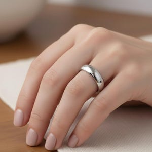 May include: A polished, silver-colored ring with a smooth, rounded design. The ring is worn on a finger, reflecting light and showcasing its simple, elegant form. The ring's width is approximately 0.25 inches.