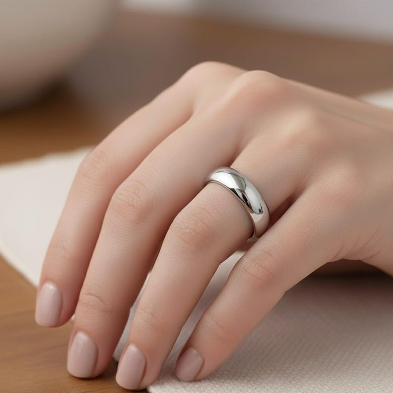 May include: A polished, silver-colored ring with a smooth, rounded design. The ring is worn on a finger, reflecting light and showcasing its simple, elegant form. The ring's width is approximately 0.25 inches.