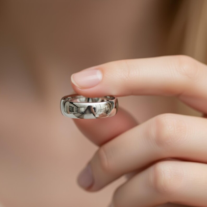 May include: A polished silver ring with a rounded band. The ring is held between two fingers, reflecting the surrounding environment. The ring's smooth surface catches the light, creating a bright, reflective appearance. A simple, elegant piece of jewelry.
