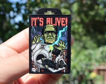 May include: A black and green enamel pin featuring a cartoon illustration of Frankenstein's monster with the text "IT'S ALIVE!"