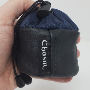 May include: A small, drawstring pouch with a navy blue top and black leather bottom. The pouch has a black cord and a black plastic toggle. The brand name "Chasm." is printed on a black label.