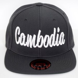 May include: A charcoal gray flat-brimmed baseball cap with the white cursive word "Cambodia" embroidered on the front. The cap has a structured crown and a curved brim. A black sticker is on the brim.