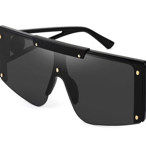 Pu&ograve; includere: Occhiali da sole neri oversize con montatura rettangolare. Gli occhiali da sole hanno lenti scure e sono accentuati da piccole borchie color oro sulla montatura. Le aste sono nere ed eleganti, offrendo un'estetica moderna.