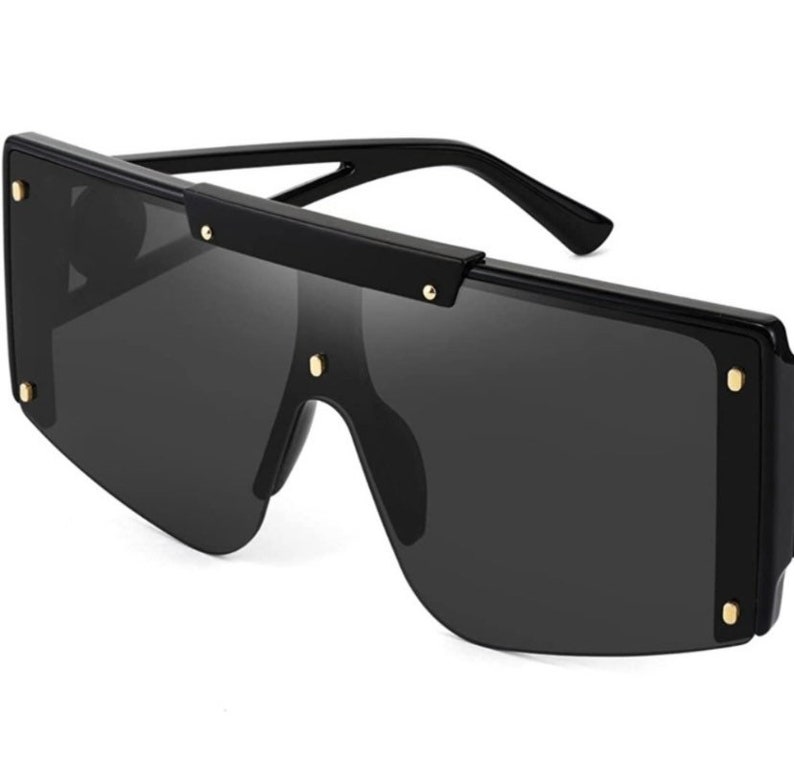 Pu&ograve; includere: Occhiali da sole neri oversize con montatura rettangolare. Gli occhiali da sole hanno lenti scure e sono accentuati da piccole borchie color oro sulla montatura. Le aste sono nere ed eleganti, offrendo un'estetica moderna.