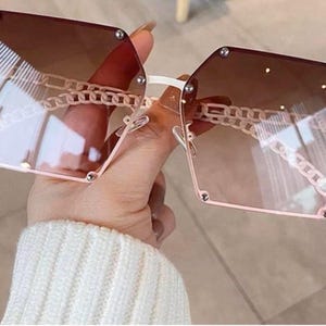 May include: Square-framed sunglasses with a gradient brown tint. The frames are a light pink color, with a chain detail connecting the lenses. The arms are white. The sunglasses are held by a person wearing a white sweater.