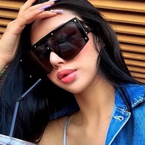 Pu&ograve; includere: Occhiali da sole neri oversize con lenti scure. Gli occhiali hanno una forma quadrata con dettagli dorati sugli angoli. L'immagine mostra un primo piano degli occhiali da sole.