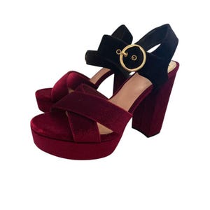 May include: Pair of burgundy velvet platform sandals with a black ankle strap and gold buckle. The sandals have a thick block heel and a criss-cross strap design across the foot.