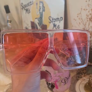 May include: Oversized square sunglasses with transparent frames and orange-tinted lenses. The sunglasses are held up in front of a vintage sign that says "Squeeze Me" and "Stomp Me".