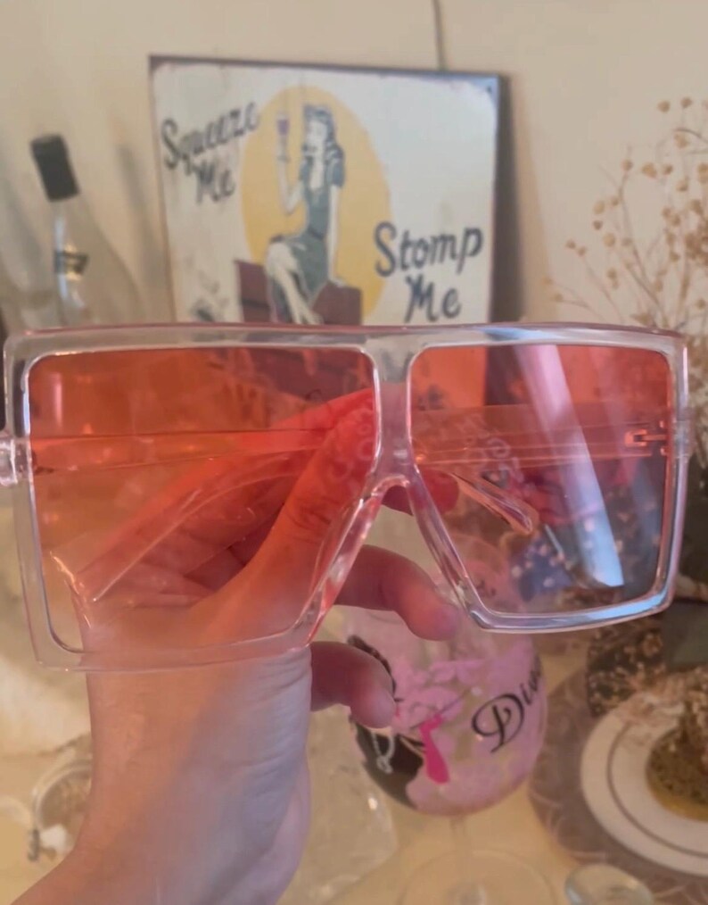 May include: Oversized square sunglasses with transparent frames and orange-tinted lenses. The sunglasses are held up in front of a vintage sign that says "Squeeze Me" and "Stomp Me".