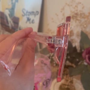 May include: Clear pink-tinted sunglasses with a rectangular frame. The temples and frame are transparent, and the lenses are a solid pink. The glasses are in a clear plastic bag.