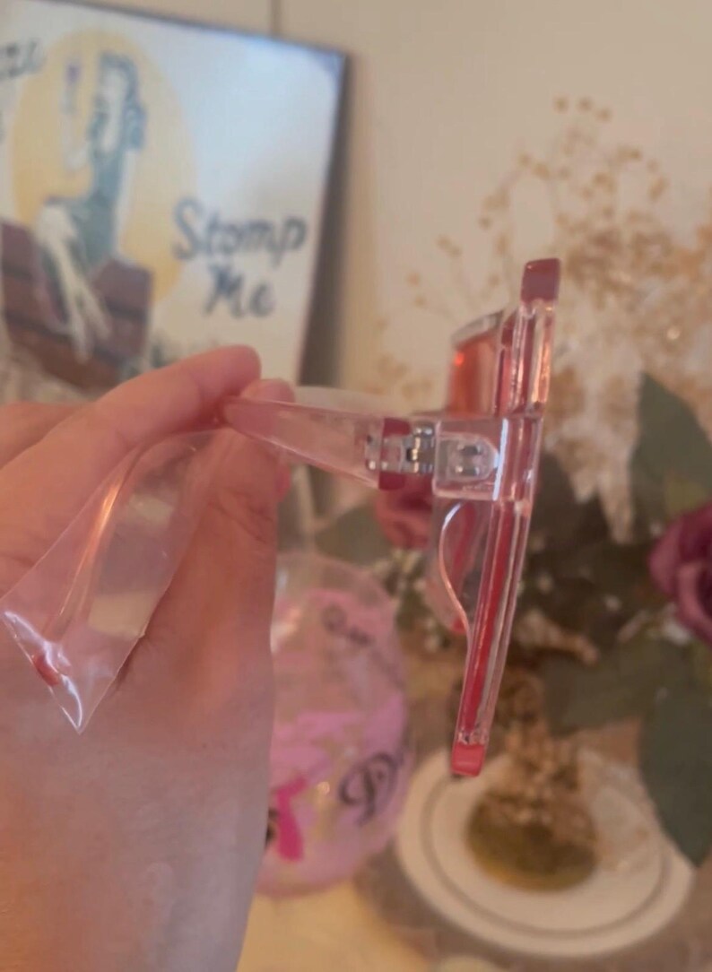 May include: Clear pink-tinted sunglasses with a rectangular frame. The temples and frame are transparent, and the lenses are a solid pink. The glasses are in a clear plastic bag.