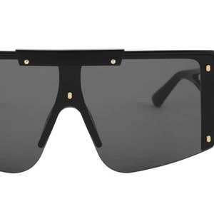 Pu&ograve; includere: Occhiali da sole neri oversize con lenti grigio scuro. Gli occhiali hanno una montatura rettangolare spessa con dettagli dorati sugli angoli e sul ponte. Le aste sono sottili e nere e le lenti sono grigio scuro.