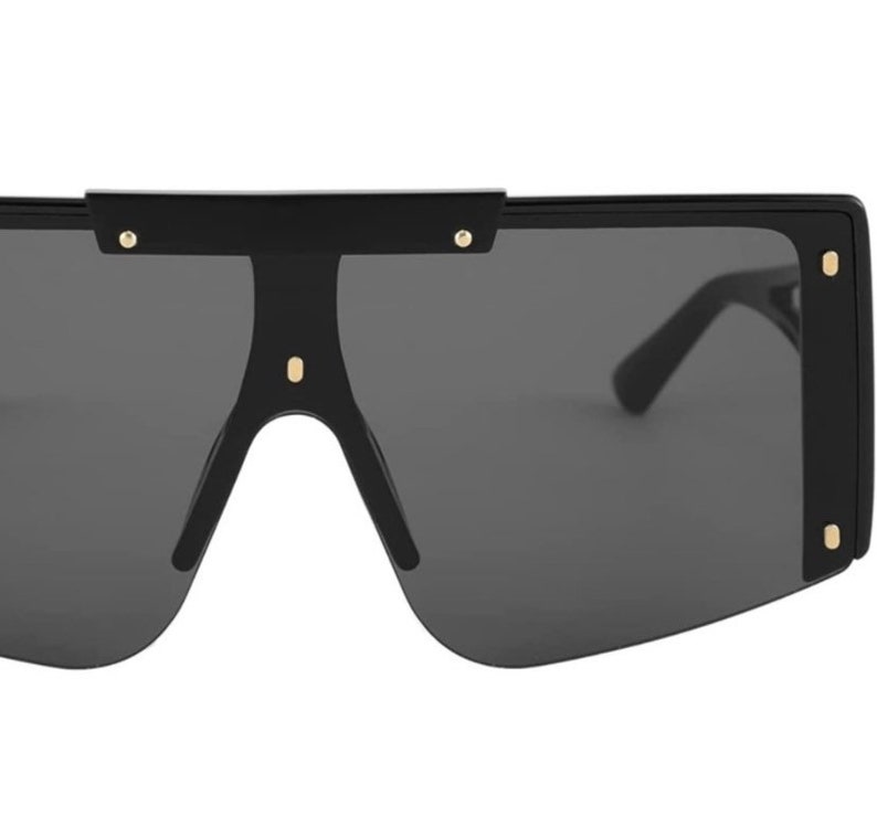 Pu&ograve; includere: Occhiali da sole neri oversize con lenti grigio scuro. Gli occhiali hanno una montatura rettangolare spessa con dettagli dorati sugli angoli e sul ponte. Le aste sono sottili e nere e le lenti sono grigio scuro.