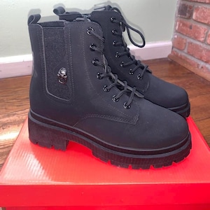 May include: A pair of black combat boots with a lug sole. The boots have a lace-up design and a small skull charm on the side. They are sitting on a red box.