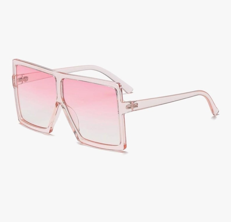 May include: Oversized square sunglasses with a pink gradient lens. The frames and arms are a translucent pink. These fashion sunglasses offer a bold, modern look.