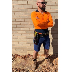 May include: A man wearing a bright orange long-sleeved shirt, blue shorts, and black work boots. He is wearing a black belt with a tool holder attached to it. The man is standing in front of a brick wall.