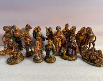 May include: A collection of ceramic nativity figurines, including figures of people, camels, and other animals. The figurines are painted in shades of brown, orange, and green, and are arranged on a white surface.
